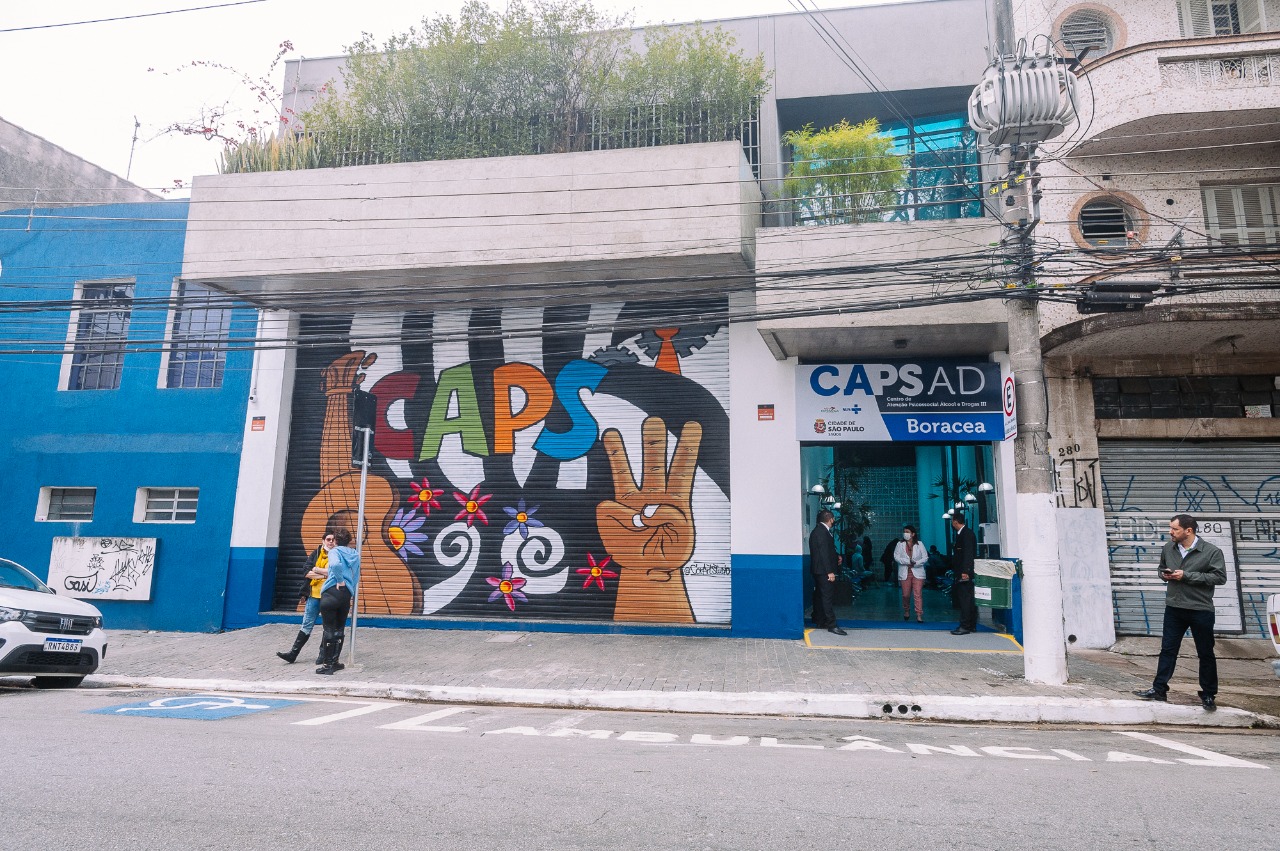 A imagem mostra a faixada do Caps. Na parede há uma arte com flores e letras coloridas. Na entrada do Caps há três pessoas.
